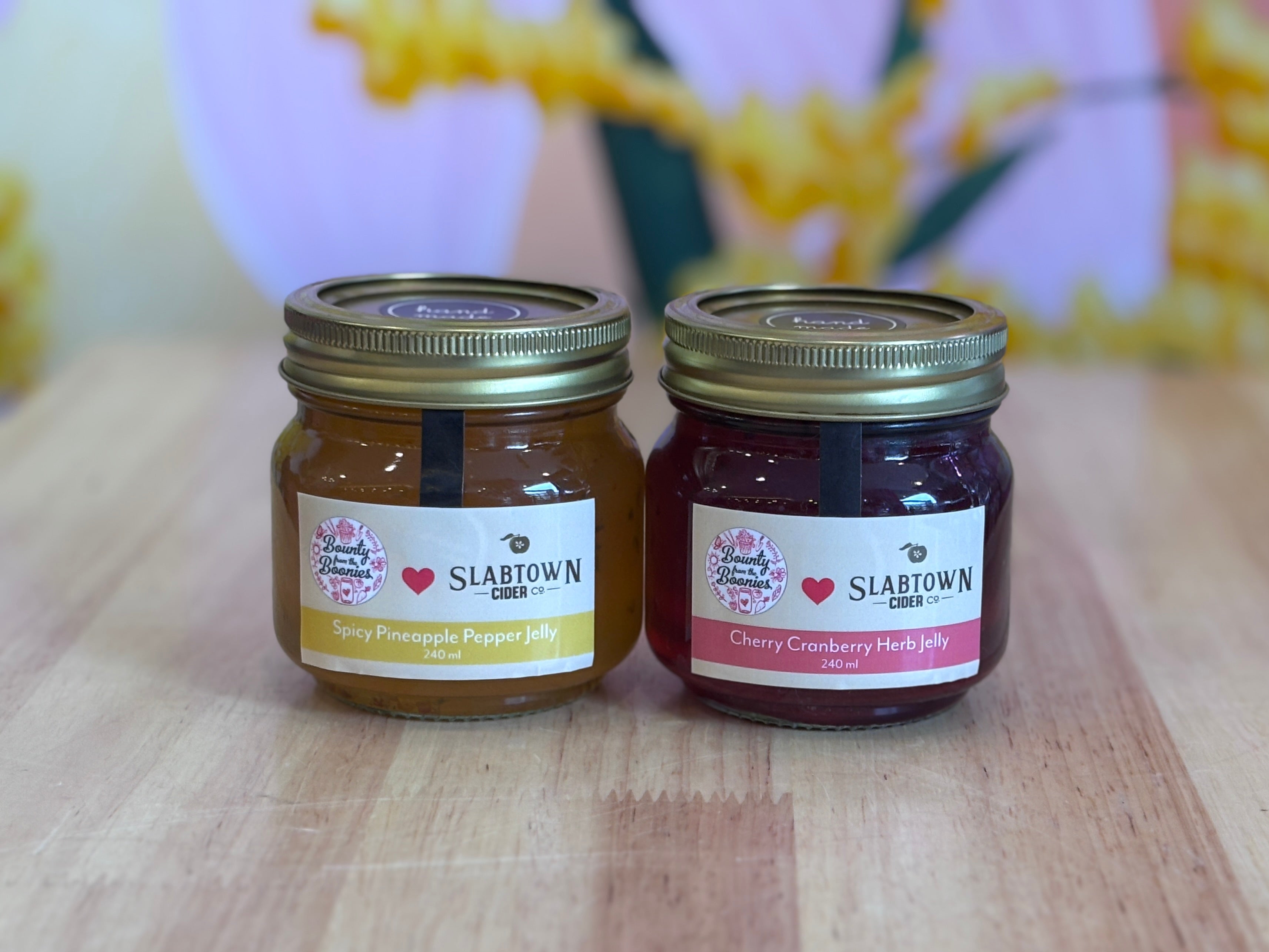 Two jars of Slabtown preserves on a wooden surface with a blurred floral background.